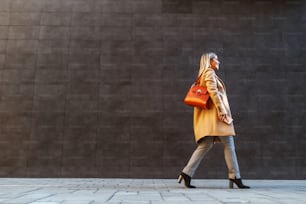 Side view of attractive caucasian blond woman in coat passing by gray wall outside.