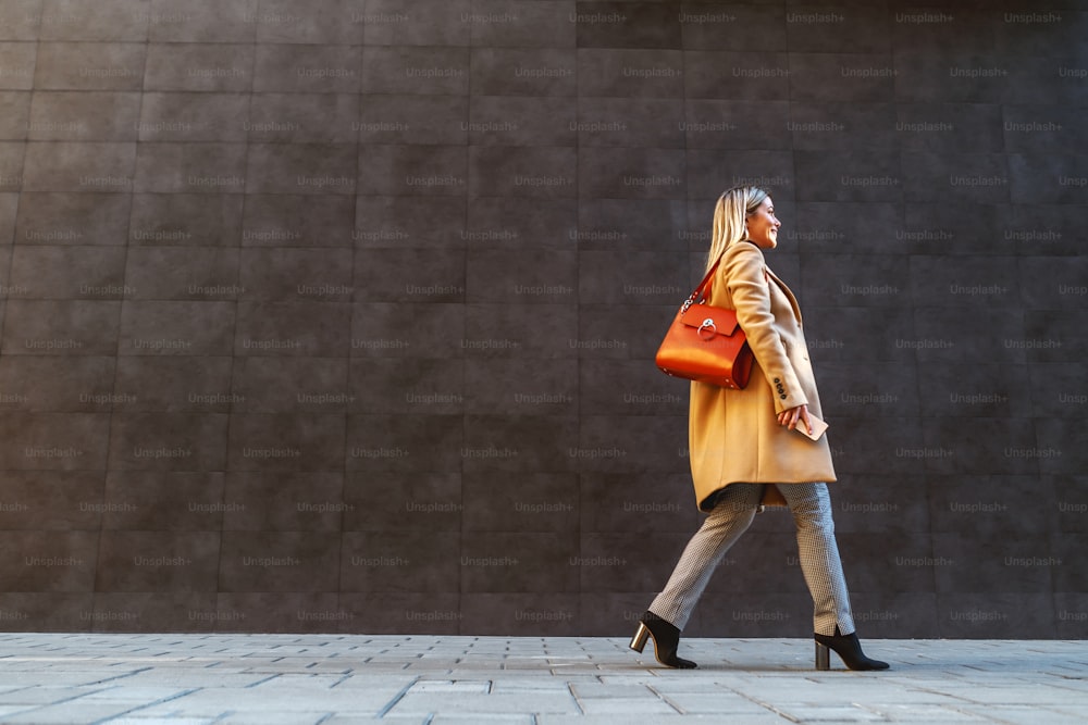 Side view of attractive caucasian blond woman in coat passing by gray wall outside.