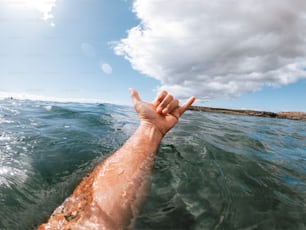 Manos del hombre en el signo del surf hallo fuera del agua azul del océano con la costa y el cielo agradable en el fondo - concepto de la gente y las vacaciones de verano