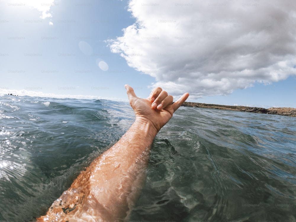 Manos del hombre en el signo del surf hallo fuera del agua azul del océano con la costa y el cielo agradable en el fondo - concepto de la gente y las vacaciones de verano