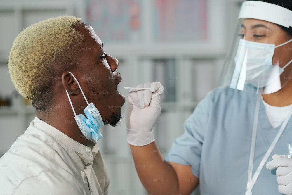 Young mixed-race female clinician in protective workwear standing in front of African man with open mouth while testing him for covid