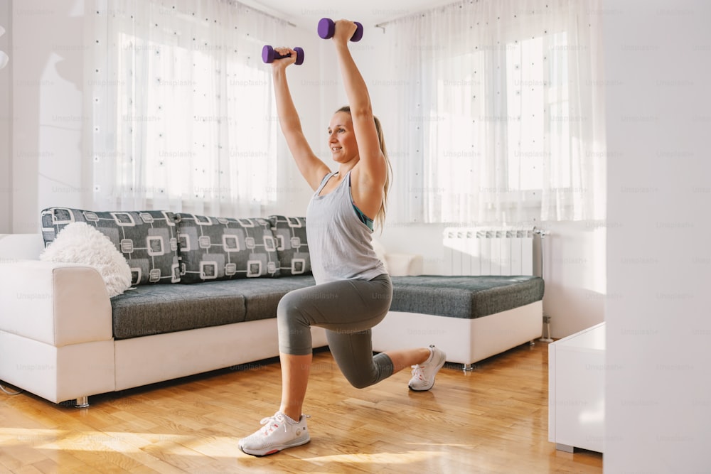 Sportswoman in excellent shape doing lunges while holding dumbbells at home.