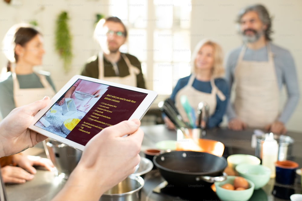 Manos de entrenador de cocina masculino con tableta mirando a través de la receta en línea de deliciosos croissants caseros contra el grupo de sus aprendices