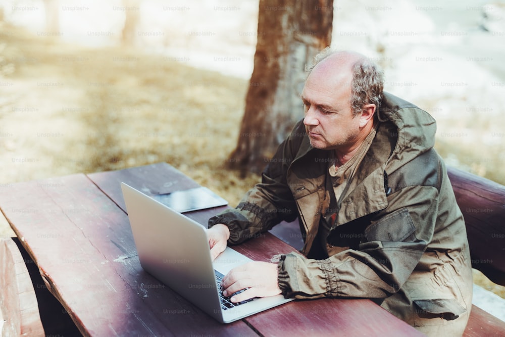 Adult male gamekeeper is sitting outdoor at a wooden table with his gadgets and using the netbook; the senior hunter in tarpaulin coverall is working on the laptop while sitting outdoors on the bench