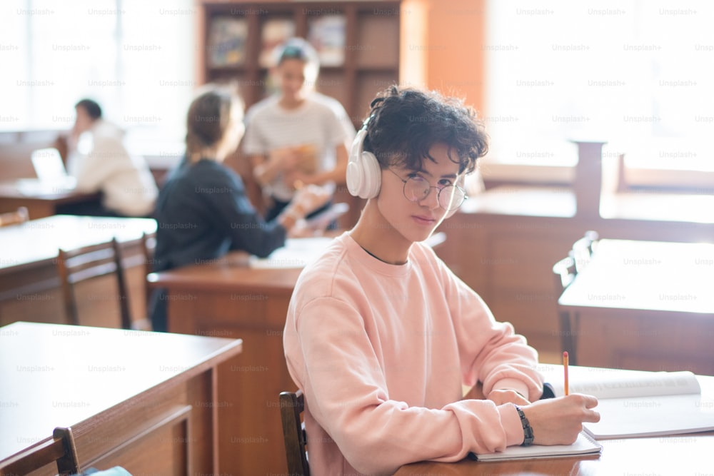 Youthful clever student of college looking at you through eyeglasses while listening to music in headphones and working by desk