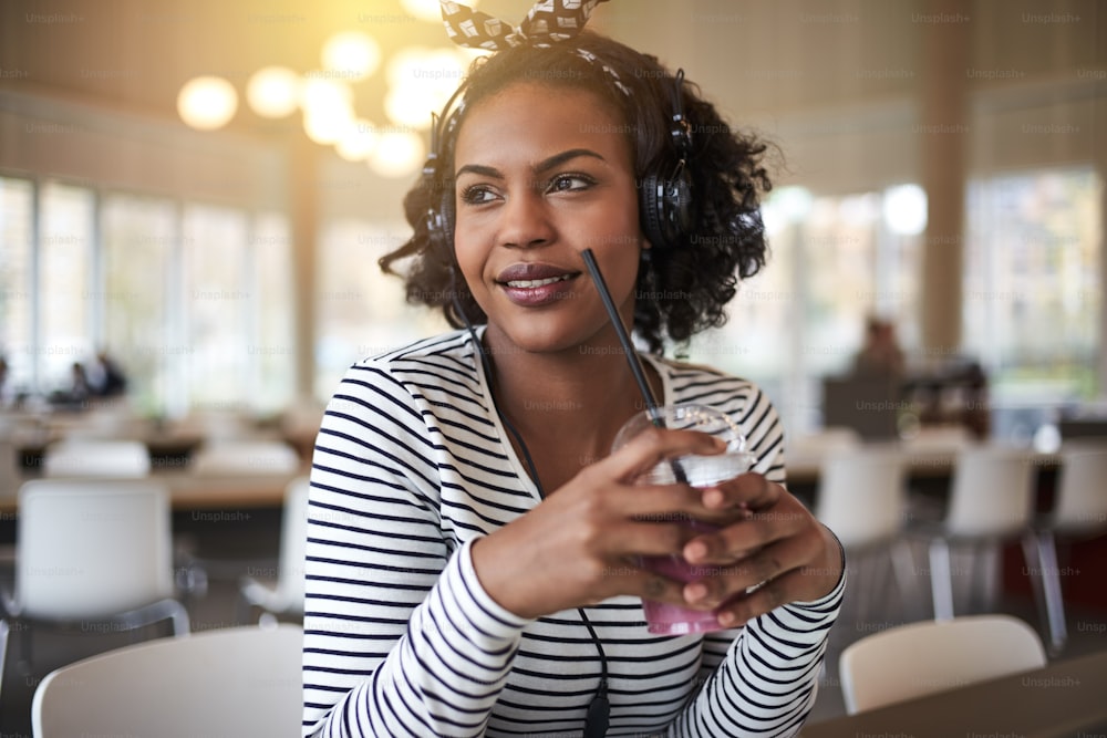 Lächelnde junge afrikanische Studentin, die über ihre Zukunft nachdenkt, während sie auf dem Campus sitzt, einen Smoothie trinkt und Musik über Kopfhörer hört