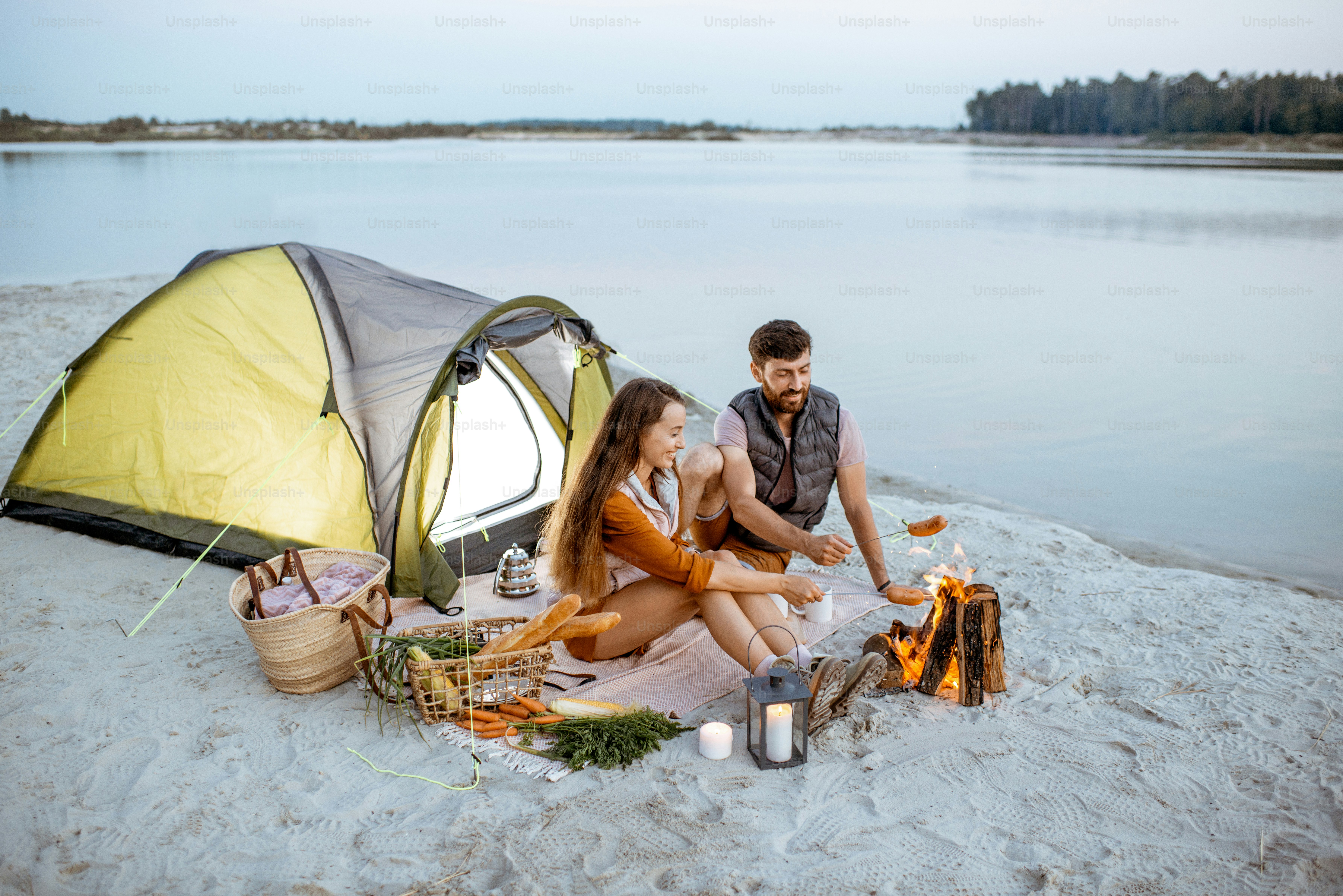 Junges und fröhliches Paar sitzt am Kamin, kocht Würstchen, picknickt abends auf dem Campingplatz am Strand