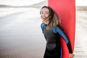 Retrato de una mujer joven y alegre en traje de neopreno de pie con una tabla de surf roja en la playa del océano durante una puesta de sol. Concepto de deporte acuático y estilo de vida activo