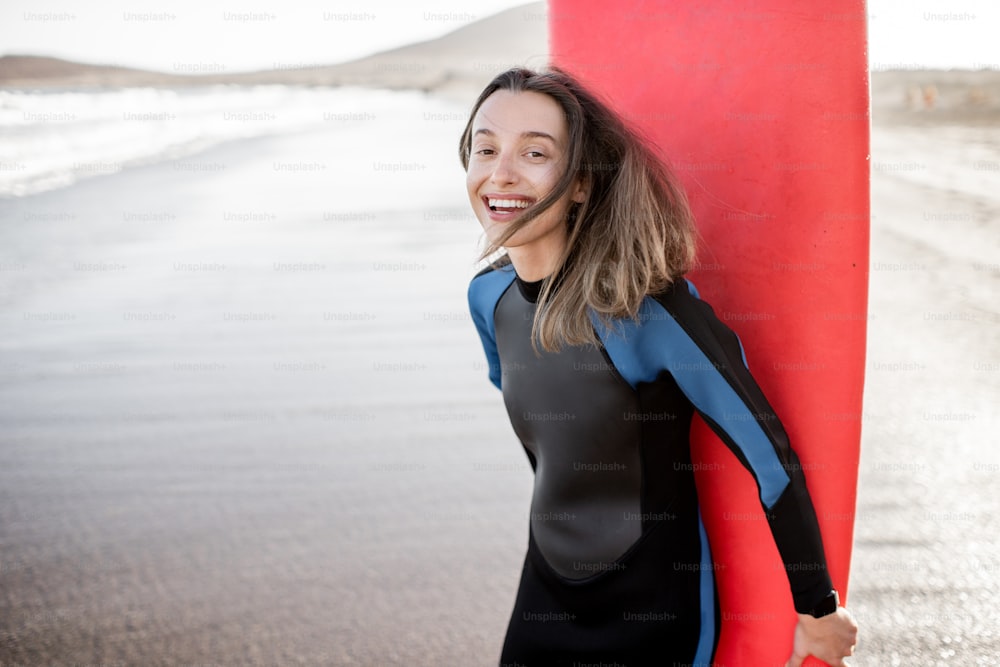 Portrait of a young joyful woman in wetsuit standing with red surfboard on the ocean beach during a sunset. Water sport and active lifestyle concept
