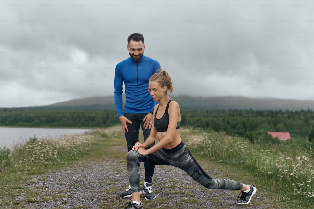 Two mid age man and woman exercising outdoor, warming up arm muscules, in summer, on gloomy day, standing at the road with scenic view, doing warm up stretching, wearing blue jacket, he has a beard