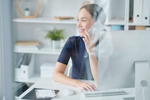 Happy young elegant woman holding smartphone by ear while sitting by desk in front of computer monitor