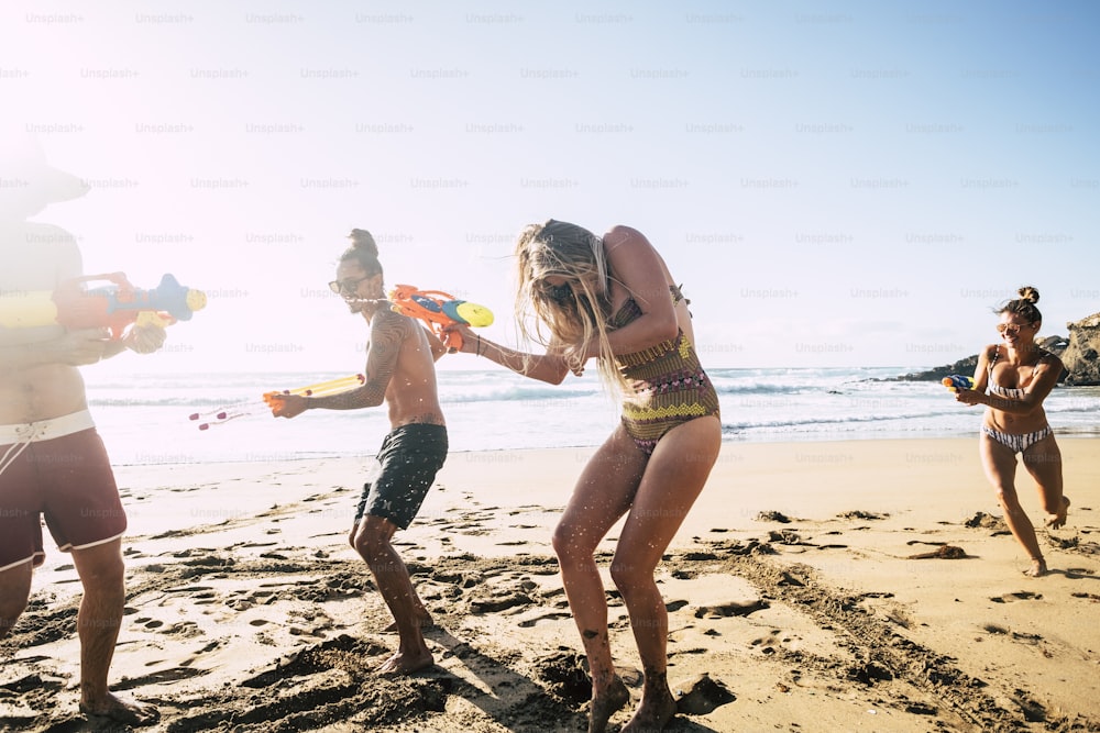 Pessoas se divertindo juntas em amizade na praia brincando com armas de água de biquíni sob o sol quente do verão - loucura e amigos - imagem brilhante homens e mulheres bonitos com o mar ao fundo