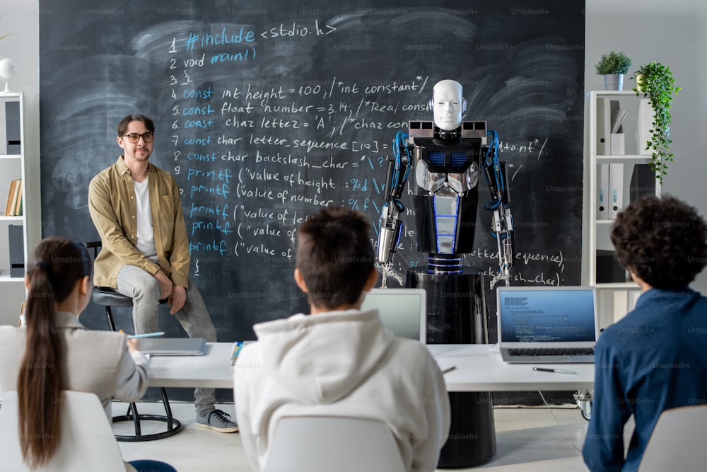 Group of teenage students looking at robot standing by blackboard and describing his technical characteristics to teacher at seminar
