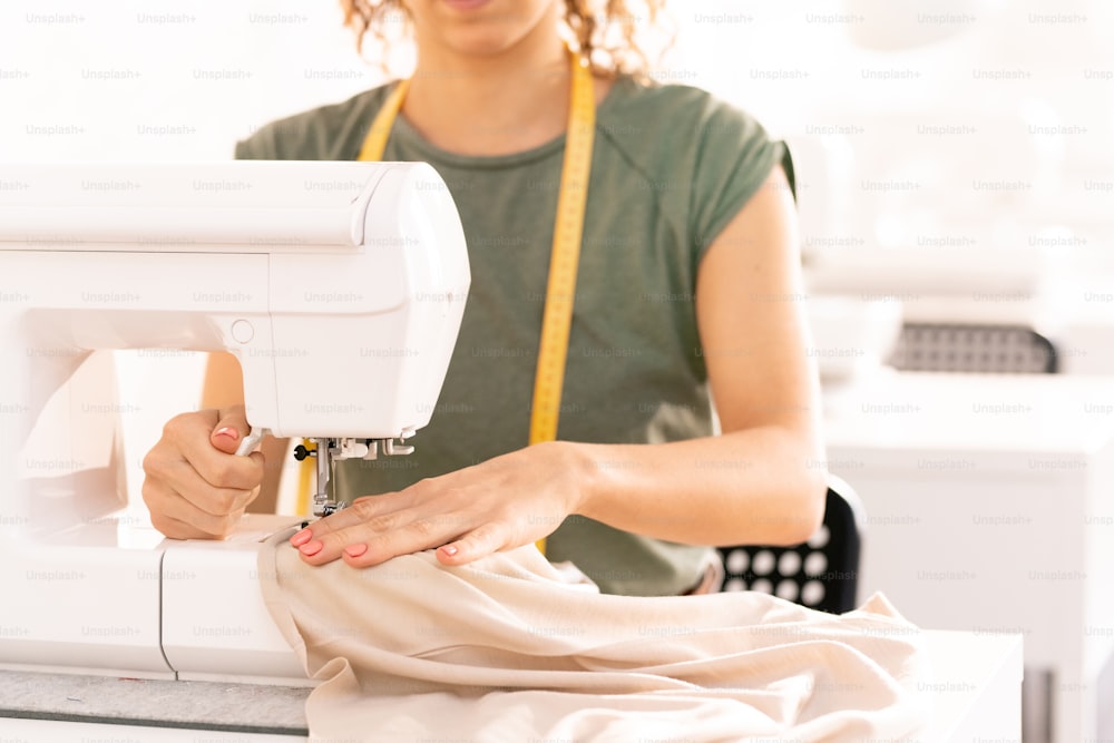 Costurera profesional sentada junto a la máquina de coser mientras trabaja en la nueva colección de temporada en el taller