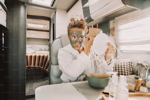 Caucasian woman making beauty skin treatment using cream and mirror sitting and relaxing inside a camper van in travel vanlife lifestyle. Adult female people have care and use mask for aging