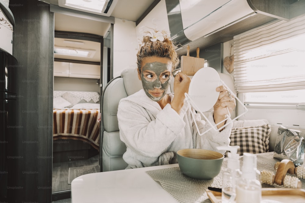 Caucasian woman making beauty skin treatment using cream and mirror sitting and relaxing inside a camper van in travel vanlife lifestyle. Adult female people have care and use mask for aging