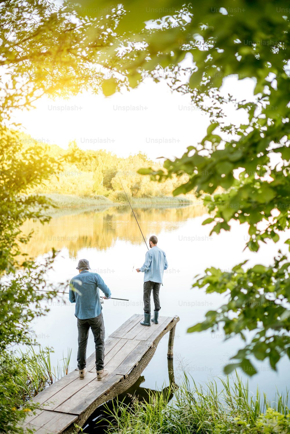 Dois amigos do sexo masculino pescando juntos de pé no cais de madeira durante a luz da manhã no belo lago