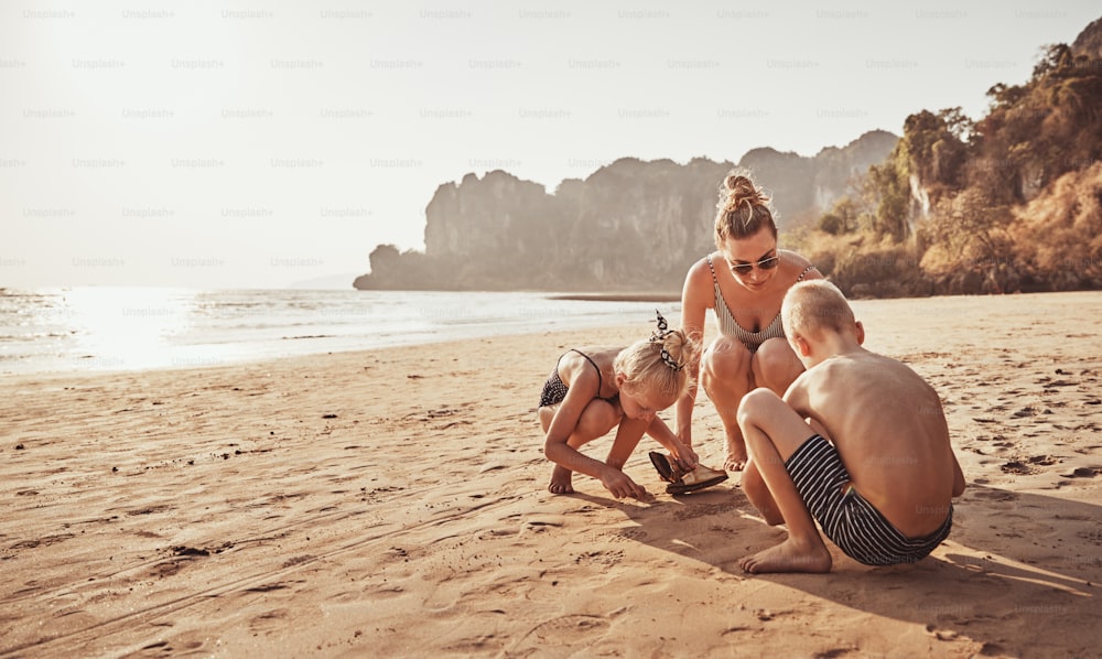 Madre sorridente e i suoi due bambini che giocano nella sabbia durante una vacanza insieme in spiaggia