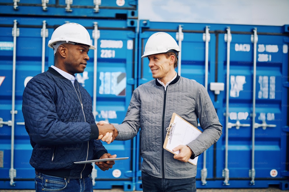 Two smiling engineers wearing hardhats standing by shipping containers on a commercial dock shaking hands together
