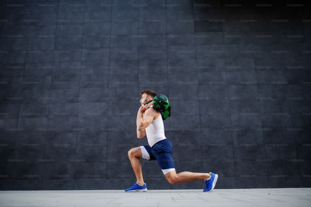 Side view of handsome muscular bearded caucasian man in shorts and t-shirt holding training bag while doing lunges.