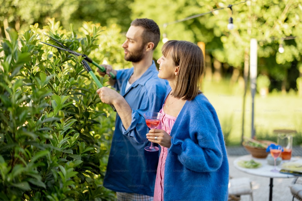 Lovely couple spend summer time at backyard of their country house on nature. Man pruning green bushes, woman have a drink and talk with husband