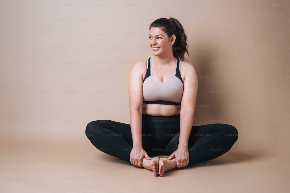 Self loving young woman plus size in sport wear doing stretching on beige background, body love
