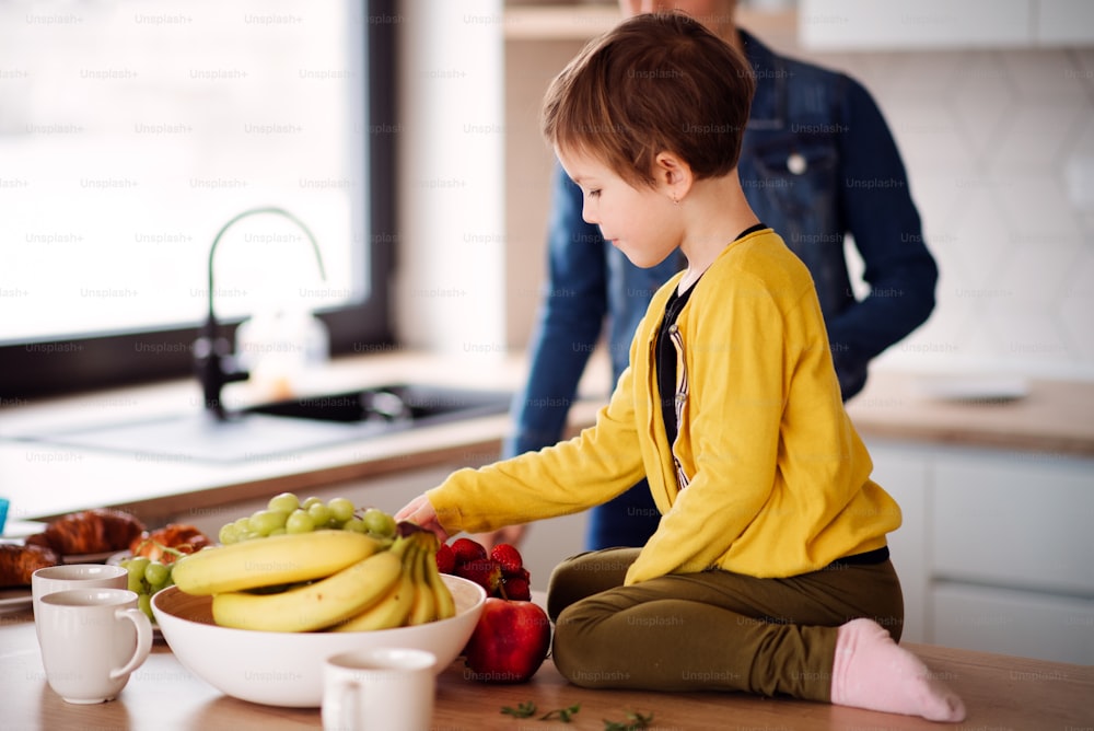 A small girl with unrecognizable mother sitting on the counter in a kitchen, eating fruit.