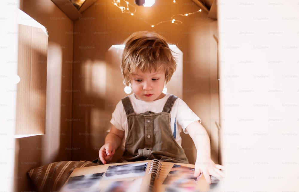 Um menino brincando dentro de casa de papelão em casa, olhando para o álbum de fotos.