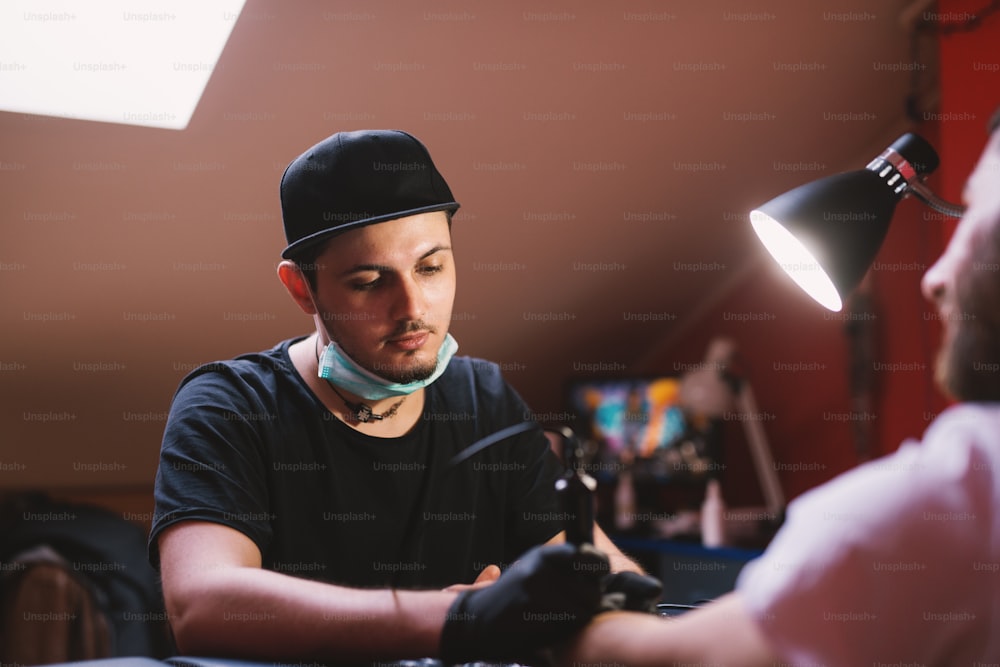 Young focused tattoo artist is inking customers arm carefully in his shop.