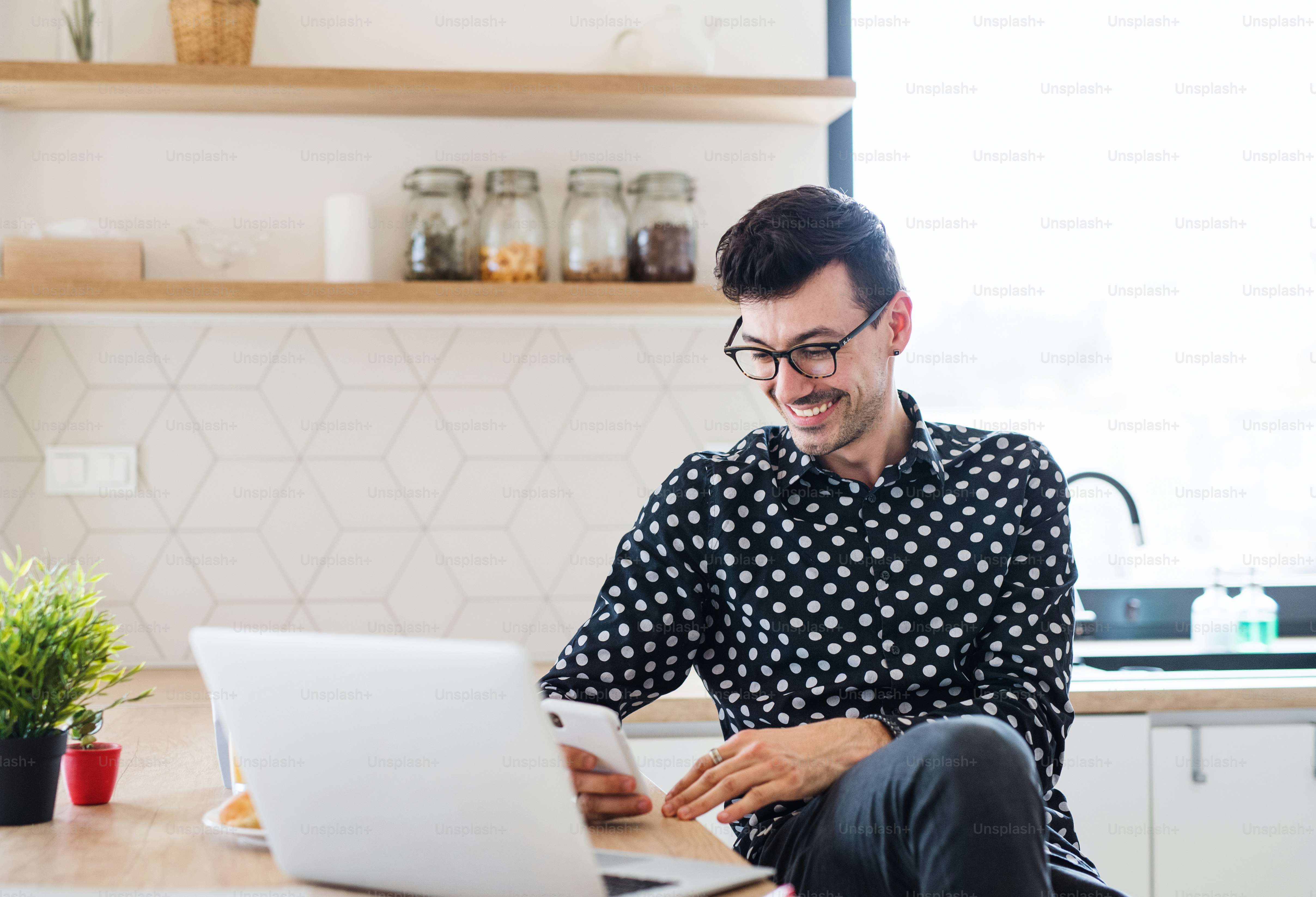 Junger Mann mit Laptop und Smartphone sitzt in der Küche und schreibt SMS. Ein Home-Office-Konzept.