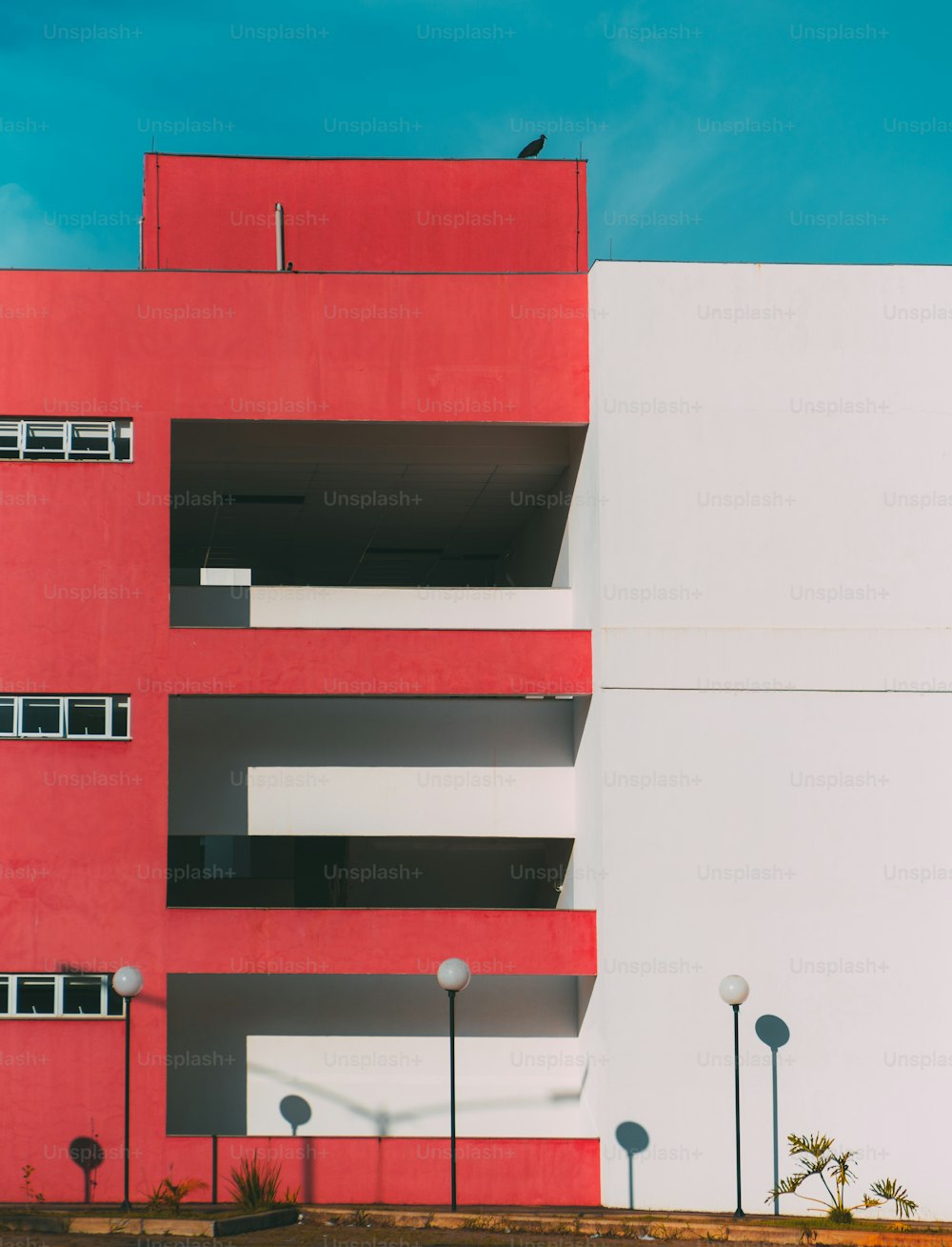 Die Fassade des modernen Gebäudes ist zweigeteilt: Ein Teil der Fassade ist rot und hat Balkone und Fenster, ein anderer Teil ist weiß; Laternen unten, ein riesiger Vogel auf dem Dach, minimalistische Geometrie