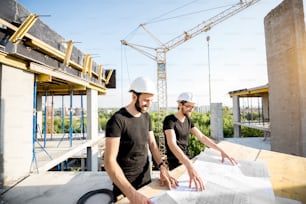 黒いTシャツを着て防護用のハルハットを着た2人の作業員が、屋外の建設現場で図面を描いている