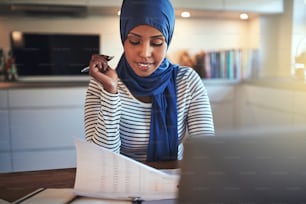 히잡을 쓴 젊은 아랍 여성이 부엌 테이블에 앉아 문서를 읽고 노트북으로 작업하고 있다