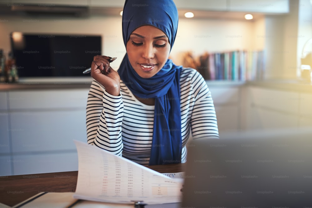 Young Arabic woman wearing a hijab reading through documents and working on a laptop while sitting at a table in her kitchen