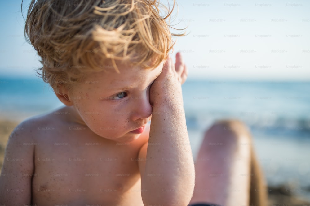 A close-up of small blond toddler boy on beach on summer holiday. Copy space.