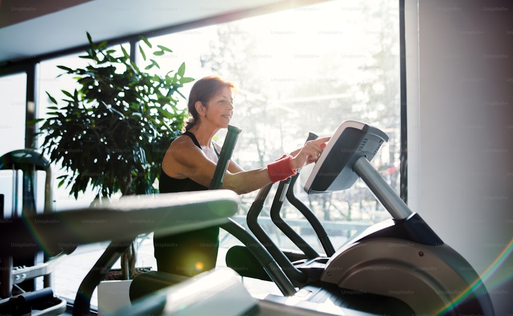 A cheerful female senior in gym doing cardio workout, exercising on stationary bicycle.