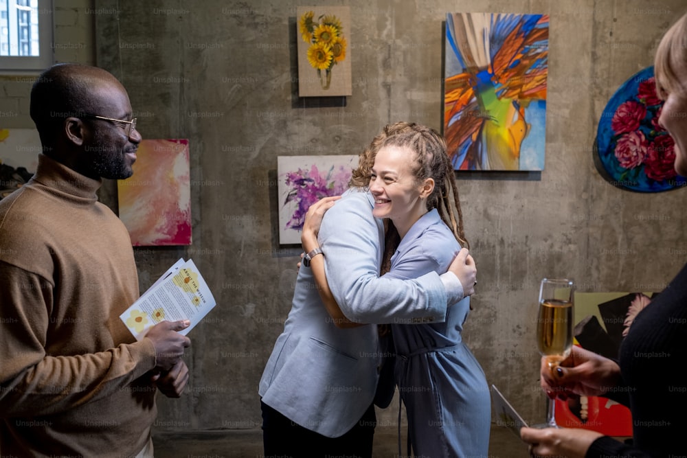Glückliche junge Frau, die einen modernen Künstler umarmt, während sie mit einer Sammlung seiner Gemälde in der Kunstgalerie an der Wand steht
