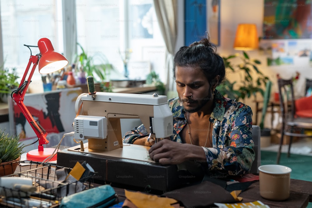 Retrato do jovem indiano criativo sentado à mesa na sala de estar em casa usando a máquina de costura para fazer algumas roupas novas