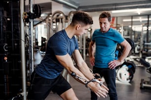 A senior man with a young male trainer doing strength workout exercise in gym.