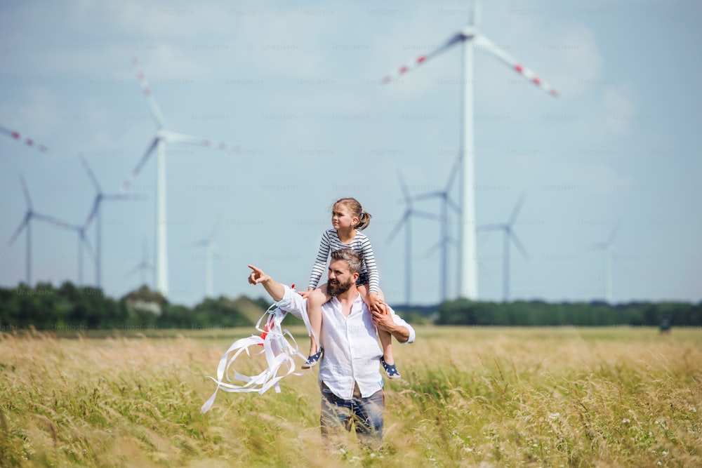 Mature father with small daughter walking on field on wind farm, giving her piggyback ride.