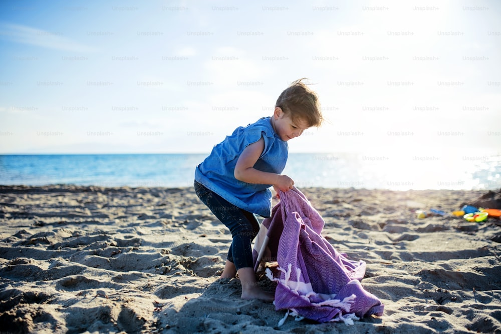 A portrait of happy small girl standing outdoors on sand beach, playing.