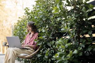 Mulher jovem trabalha no computador portátil enquanto sentada relaxada na cadeira no fundo de arbustos verdes no quintal. Conceito de trabalho remoto em ambiente aconchegante