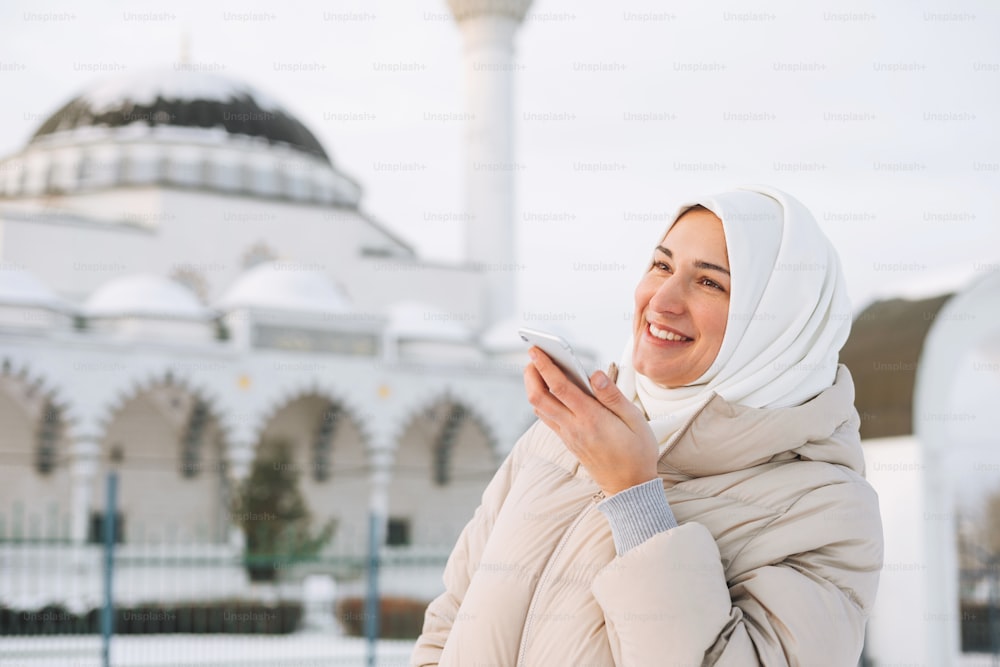 Hermosa joven musulmana sonriente con pañuelo en la cabeza con ropa ligera usando el móvil contra el fondo de la mezquita en la temporada de invierno