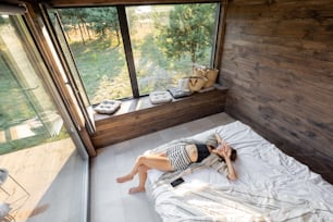 La femme se réveille dans une maison de campagne ou un hôtel avec des fenêtres panoramiques dans la forêt de pins, allongée sur le lit et bâille. Bonjour et loisirs sur le concept de nature. Vue d’en haut.