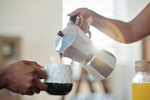 Hand einer jungen afrikanischen Frau, die Kaffee oder Tee in den Becher ihres Mannes während des Frühstücks mit einer Flasche Orangensaft in der Nähe gießt