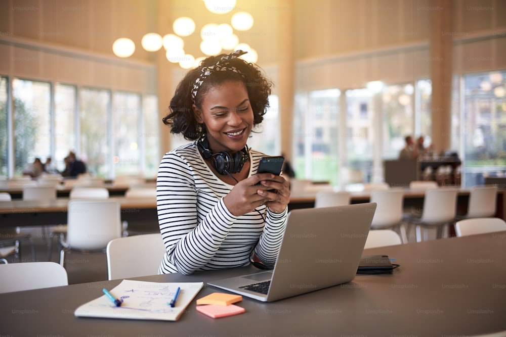 Joven estudiante universitaria africana sonriendo mientras está sentada en el campus enviando un mensaje de texto en su teléfono celular y usando una computadora portátil