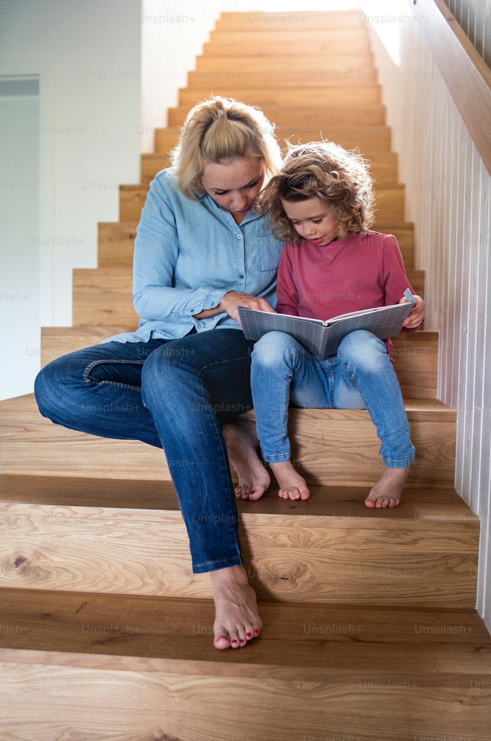 Une vue de face d’une jolie petite fille avec sa mère à l’intérieur à la maison, assise et lisant dans l’escalier.