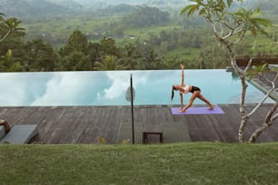 Bella ragazza che fa yoga a bordo piscina al mattino a Bali, Indonesia. Giovane donna magra in abbigliamento sportivo in piedi in posa triangolare sul tappetino sportivo vicino alla piscina a sfioro contro il paesaggio tropicale.