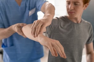 Hands of mature clinician in blue uniform supporting bent arm of young patient during physiotherapeutic training in rehabilitation clinics