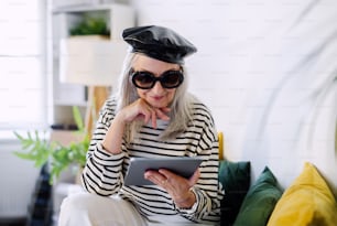 Portrait of senior woman with black beret sitting indoors at home, using tablet.